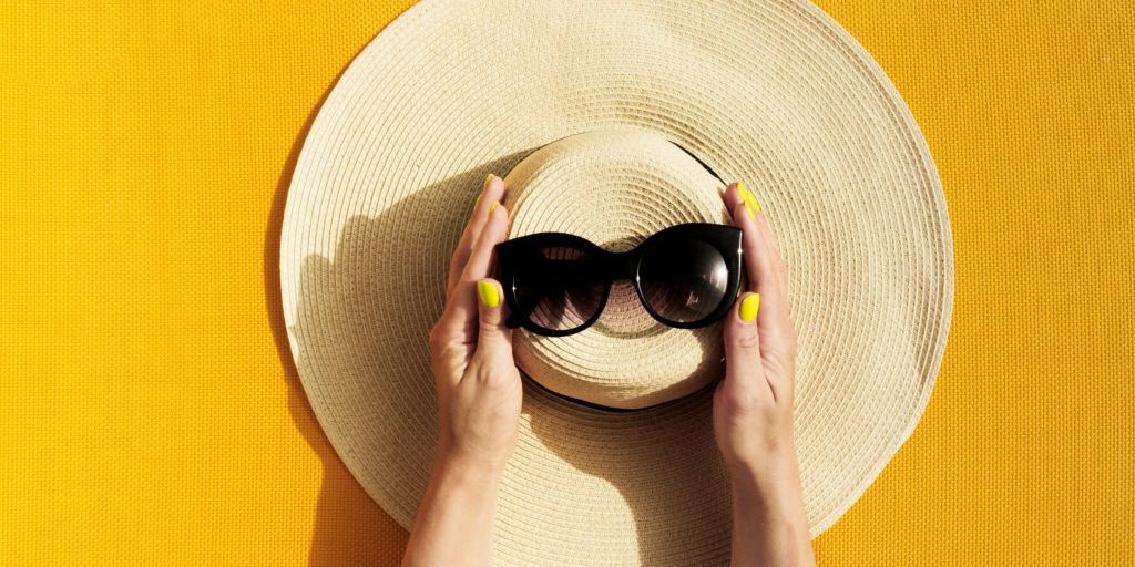 Hands of young girl holding straw hat and sunglasses on vibrant yellow background. Top View. Summer Travel Vacation Concept.