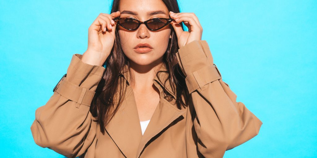 Portrait of beautiful caucasian brunette woman model in brown overcoat and sunglasses.Girl posing in studio near blue wall Portrait of beautiful caucasian brunette woman model in brown overcoat and sunglasses.Girl posing in studio near blue wall